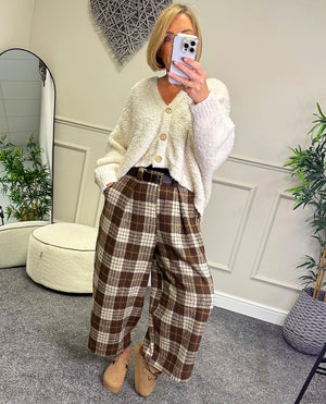 Woman taking a mirror selfie wearing a white cardigan and plaid pants in a room with plants and decor.