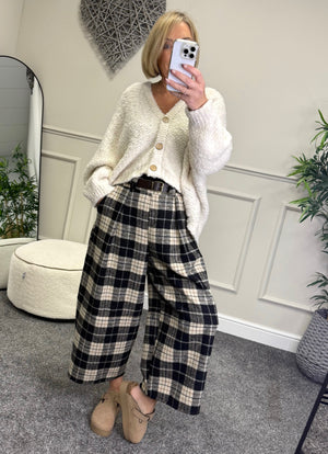 Person wearing a white cardigan, beige top, plaid pants, and beige shoes in a room with decorative elements.